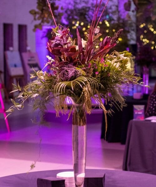 A tall, elegant glass vase filled with a vibrant floral arrangement featuring various purple and green flowers and foliage, set against a backdrop of artworks and purple ambient lighting. The table on which the vase sits is draped with a dark cloth.