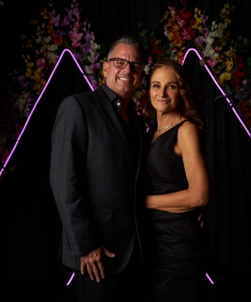 A man and a woman are standing together and smiling at the camera. They are dressed in formal attire, with the man in a suit and the woman in a black dress. The background features colorful flowers and neon lights arranged in a triangular shape.