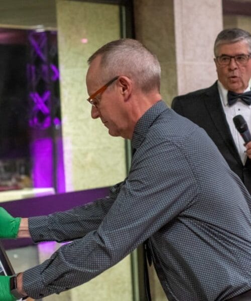 A person wearing green gloves carefully places a framed piece of art on an easel. Another person, dressed in formal attire with a bow tie, stands nearby holding a microphone. The background features large windows and colorful lights.