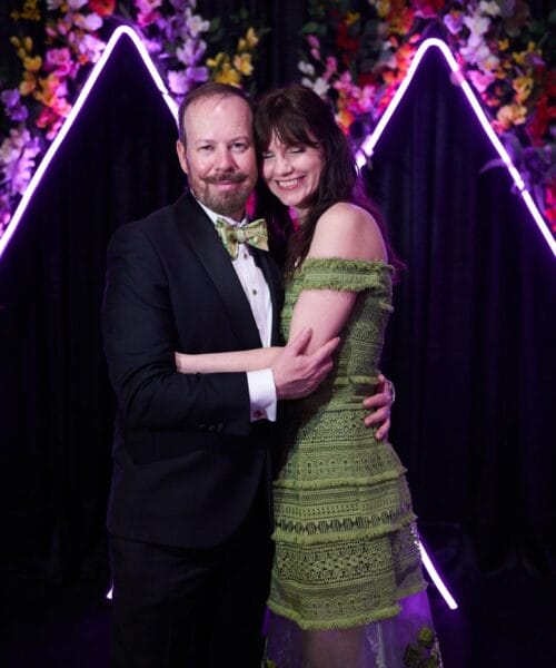 A couple dressed in formal attire poses in front of a colorful floral backdrop with purple neon lights forming triangular shapes. The man wears a black tuxedo with a patterned bow tie, and the woman wears a green lace dress. They smile warmly at the camera.