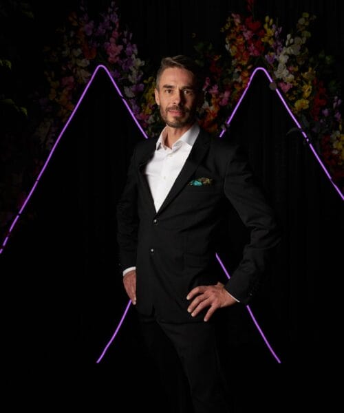 A man in a formal black suit with a white shirt stands confidently with one hand on his hip. The background features two neon purple zigzag lines and an arrangement of colorful flowers, framed by leafy green branches on each side.