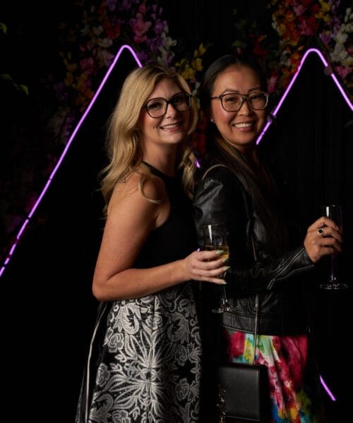 Two women stand close together, smiling at the camera in front of a dark backdrop with neon purple triangular lights and floral decorations. One wears a black dress with a patterned skirt, and the other wears glasses, a colorful dress, and a black leather jacket. Both hold drinks.