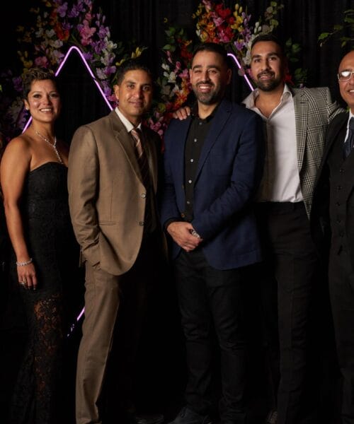 A group of six people, four men and two women, dressed in formal attire, standing together and smiling in front of a black backdrop adorned with colorful floral arrangements and neon lights. They appear to be at a celebratory or formal event.
