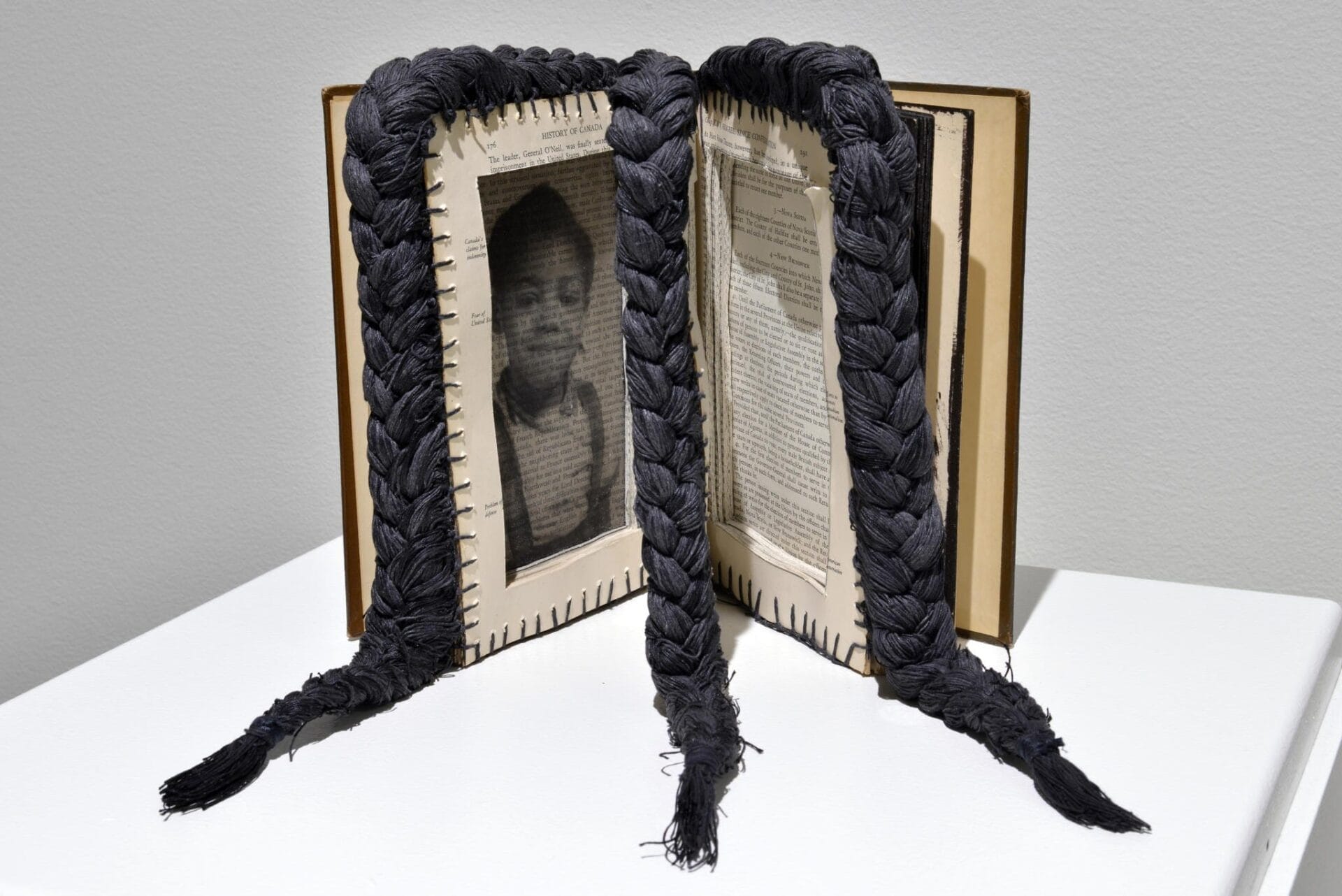 An open book stands upright with black braids extending from its pages. The left page features a black-and-white photograph of a person, while the right page has text. The braids drape down over the book and onto the surface.
