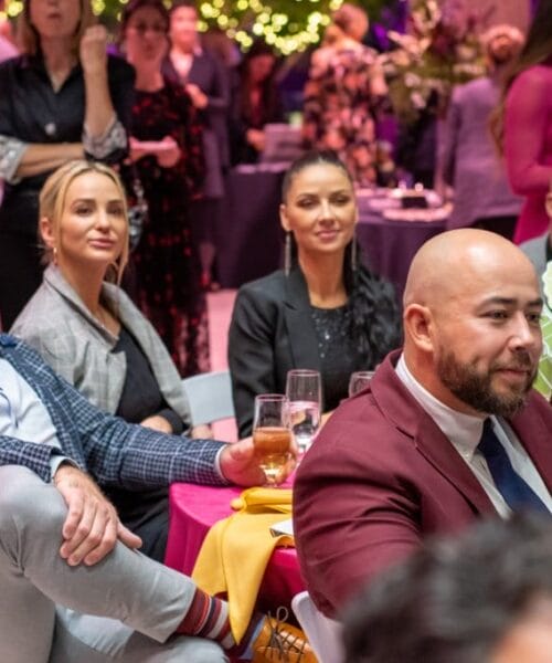 A group of people seated at a table during a lively event. They are dressed in formal attire and appear to be attentively listening to someone off-camera. The background showcases other tables with guests, festive decorations, and colorful lighting.