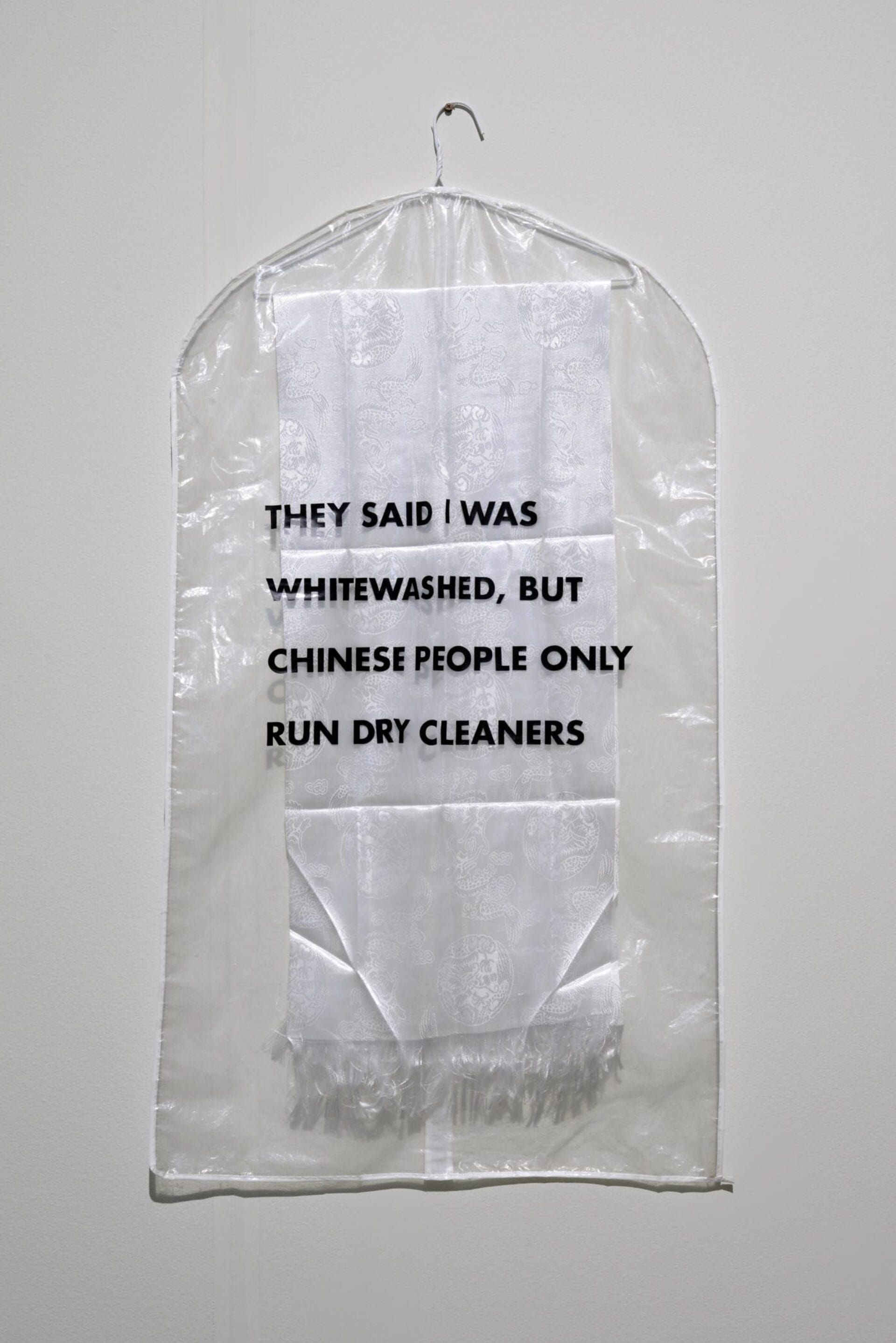 A white garment bag hangs on a wall with a white scarf inside. The text on the garment bag reads,