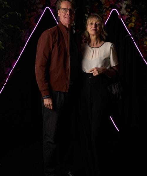 A man and woman pose together in front of a backdrop featuring neon lights in triangular shapes. The man is wearing a brown jacket and glasses, while the woman is wearing a white blouse and black pants. The background also includes flowers in various colors.