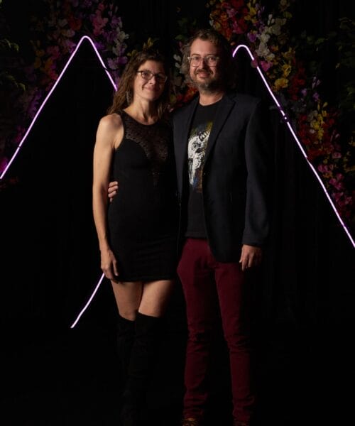 A couple posing against a dark background adorned with neon lights and colorful floral decorations. The woman is wearing a black dress and black boots, and the man is dressed in a blazer, graphic t-shirt, and maroon pants. They are smiling and standing closely together.