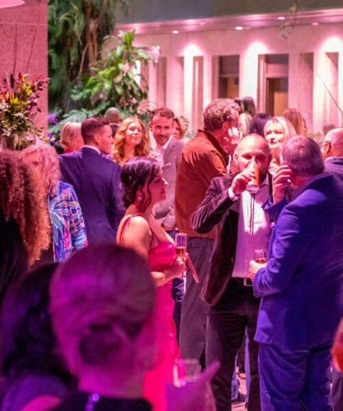 A group of people are socializing at an indoor event. The setting is well-lit with purple and pink lighting. Attendees are dressed in formal attire, holding drinks, and engaging in conversations. There are plants and modern decor elements in the background.