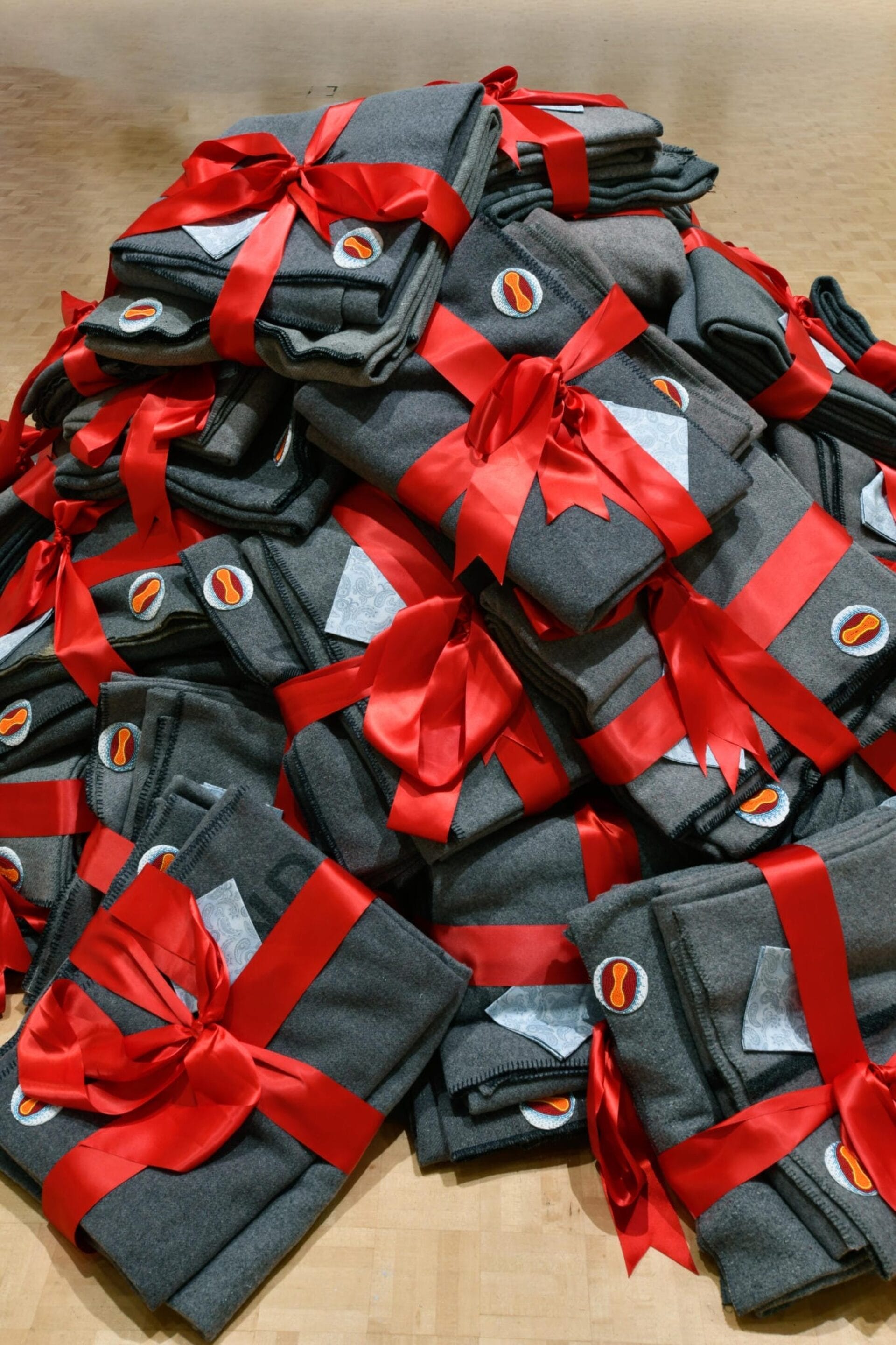 A pile of gray folded garments tied with red ribbons sits neatly stacked on a wooden surface, reminiscent of organized moving boxes. Each garment showcases a small logo patch featuring an orange oval shape with white details.