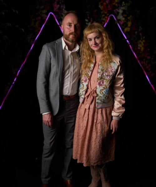 A man in a gray suit and a woman in a pink lace dress and embroidered jacket stand together in front of a dark backdrop with colorful lights and floral arrangements forming triangular shapes. They are both smiling and looking at the camera.