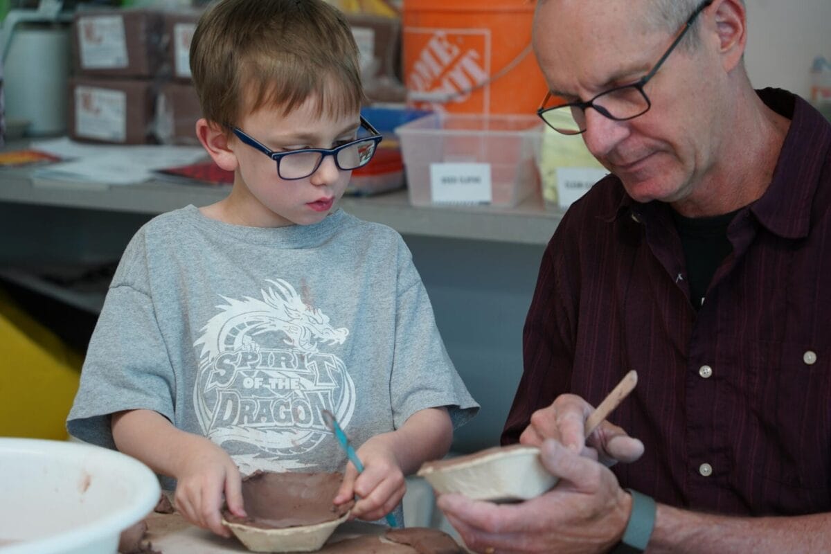 Un jeune garçon et un homme plus âgé travaillent ensemble à la poterie en argile, assis à une table. Le garçon observe attentivement l'homme qui façonne un bol avec un outil. Des fournitures artistiques et des contenants étiquetés sont visibles en arrière-plan.