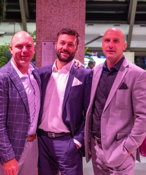 Three men are standing and smiling in a well-lit indoor setting. One wears a checkered jacket, another wears a dark suit, and the third wears a light gray suit. There are plants behind them and a woman in the background to the right.