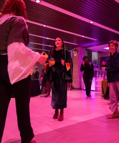 A group of people dressed in casual and semi-formal attire dance joyfully in a brightly lit room with pink and purple lighting. Some are holding drinks, and everyone appears to be enjoying the lively atmosphere.