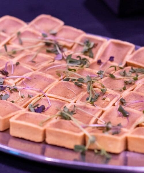 A rectangular metal tray is filled with neatly arranged small square tart shells, each garnished with fresh microgreens. The tarts are uniformly golden brown, and the tray is set against a dark background.