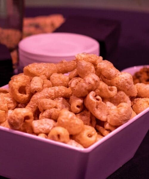 A white square bowl filled with crispy pork rinds is in focus on a dark table. In the background, there are black napkins, a white plate, and another small bowl with a different snack. The setting appears to be dimly lit.