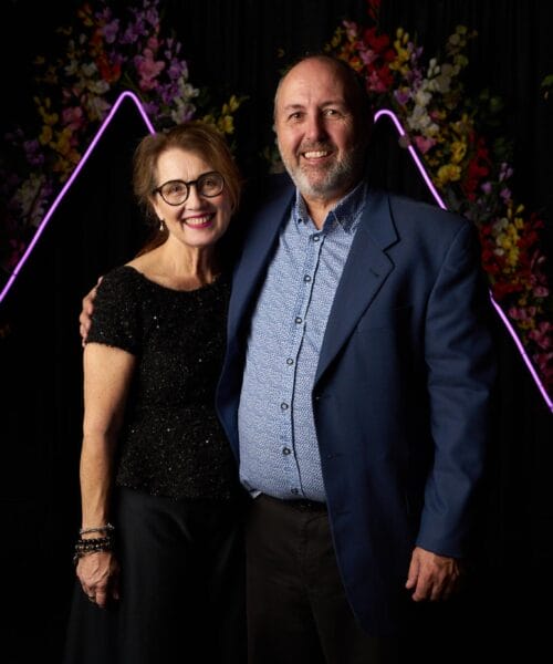 A smiling couple stands arm in arm in front of a backdrop decorated with neon purple triangular outlines and colorful flowers. The woman is wearing a black dress and glasses, while the man is wearing a blue blazer over a light blue shirt.