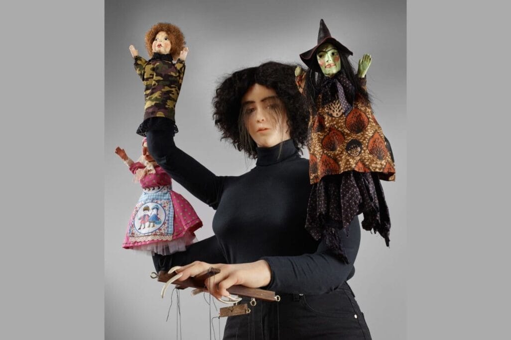 A wax figure of a lady in all black with four arms, each arm is controlling a small puppet.
