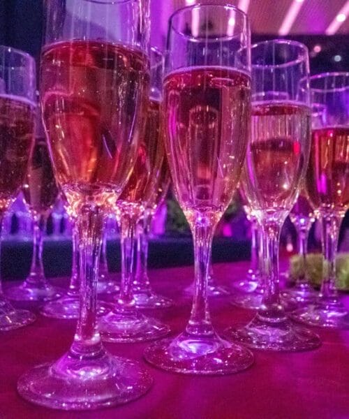 A group of filled champagne flutes on a purple-lit table, creating a festive atmosphere. The glasses contain a golden liquid, likely champagne or sparkling wine, and are arranged closely together, reflecting the vibrant lighting of the venue.