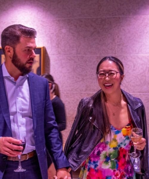 A man and a woman are attending a social event. The man is wearing a blue checkered suit and holding a glass of red wine, while the woman is wearing a colorful dress with a black leather jacket and holding a glass of white wine. They are smiling and holding hands.