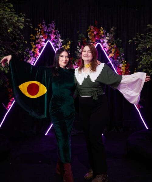 Two women stand side by side against a backdrop of vibrant flowers and neon triangle lights. The woman on the left wears a dark green dress with a large eye design on one sleeve, while the woman on the right wears a green and white outfit with wide, flowing sleeves. Both are posing with arms outstretched.