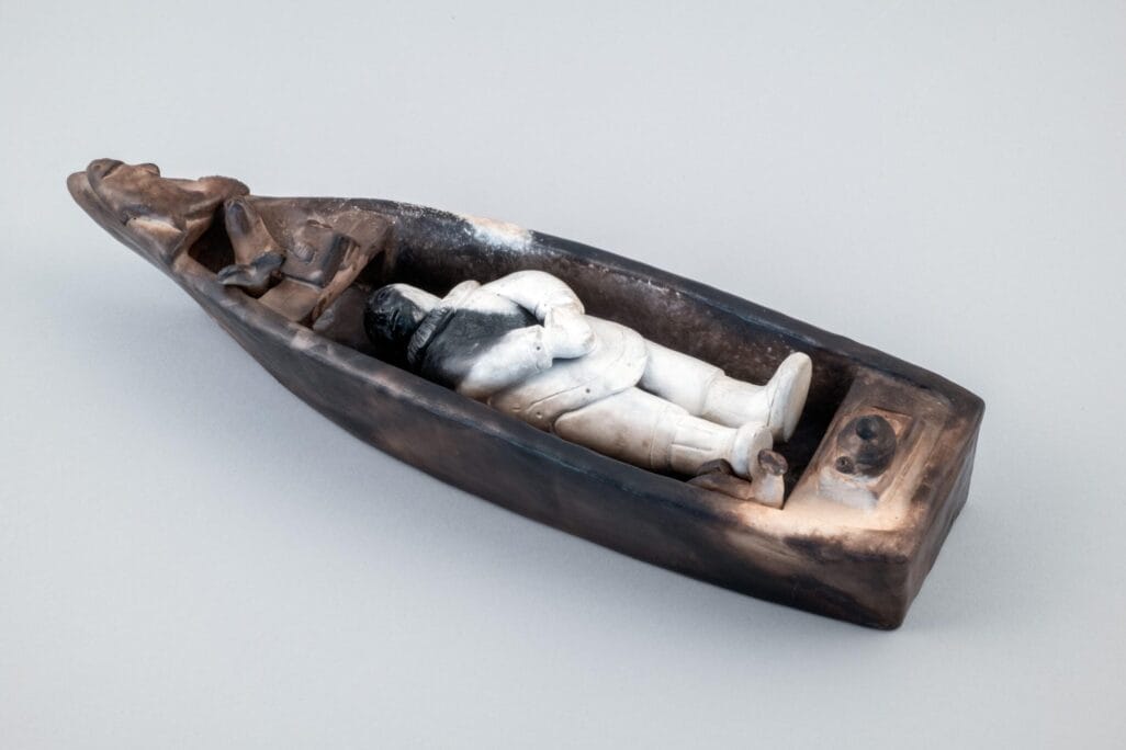 A ceramic Death Boat sculpture depicts a person lying on their back inside a narrow vessel, arms crossed over their chest, with small objects near the head and feet. The boat features dark brown and black hues with a white and gray figure.