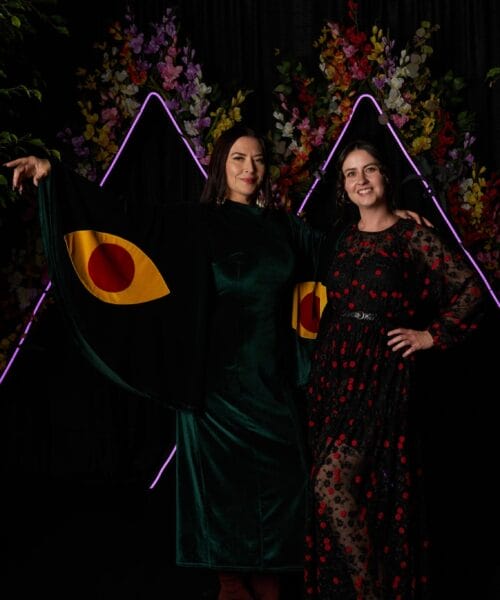 Two women pose in front of a decorative background with illuminated triangular shapes and floral arrangements. The left woman wears a green dress with large yellow eye designs on the sleeves, while the right woman wears a black dress with red floral patterns. Both are smiling.