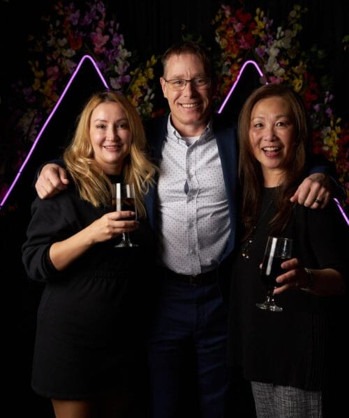 Three people, two women and one man, are posing together and smiling for a photo. Each person is holding a drink. They are standing in front of a dark backdrop with floral decorations and neon triangular lights. The atmosphere appears to be celebratory.