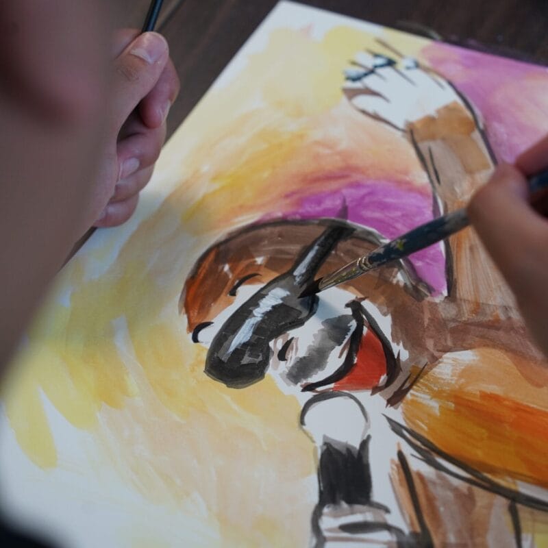 A person uses a paintbrush to add details to a colorful painting of a dog on paper, with warm tones in the background, creating unique Pop Culture Tributes through expressive art.