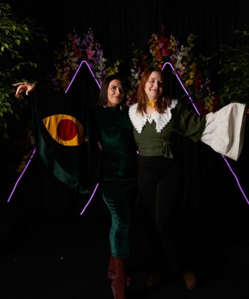 Two people stand posing with outstretched arms. One wears a black outfit with colorful sleeves featuring a yellow and red design. The other wears a green top with a white collar and flared sleeves. A backdrop of neon mountain shapes and flowers adds a festive touch.