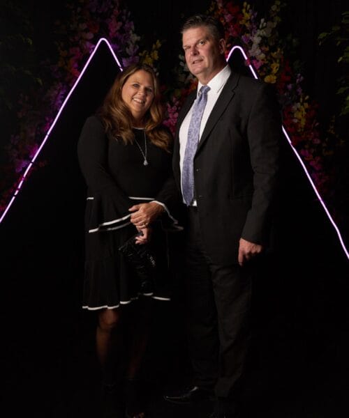 A man in a dark suit and tie stands next to a woman in a black dress with white trim. They are posing for a photo in front of a backdrop featuring neon triangle shapes and colorful flowers.