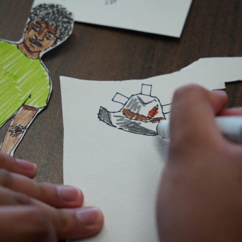 A person’s hands draw a hat on white paper with a black marker. Next to the drawing are paper cutouts, including a hand-cut, colored figure in a green shirt showing a tattoo on their forearm.