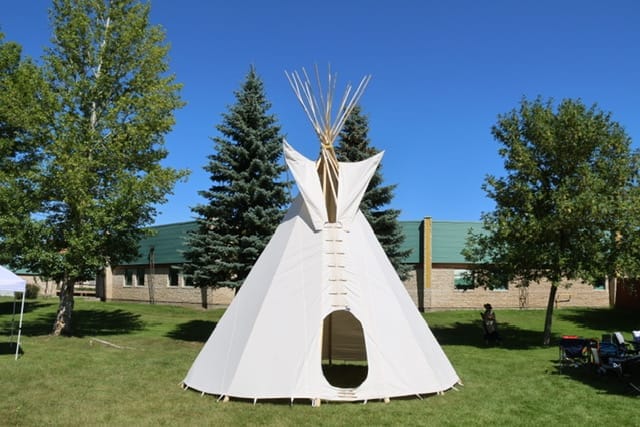 A white canvas tipi stands on a grassy lawn, surrounded by trees with a building in the background under a clear blue sky.