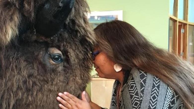 A woman with long hair and patterned clothing gently presses her forehead to a bison's forehead, creating a peaceful, intimate moment indoors.
