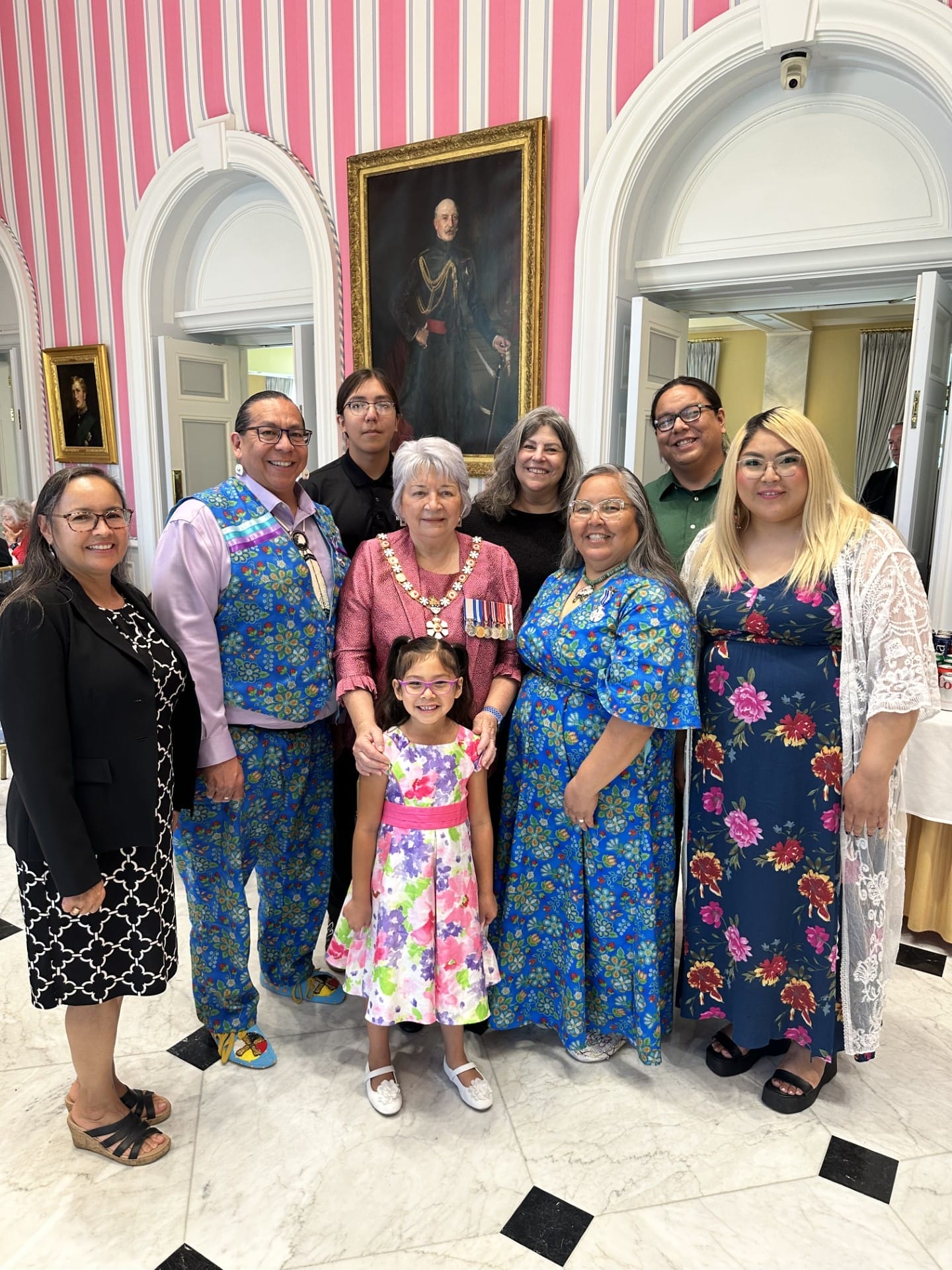 A group of nine adults and one child pose indoors, all smiling. They wear colorful, formal attire. Behind them are arched doorways, striped pink walls, and a framed portrait of a man in historical dress.