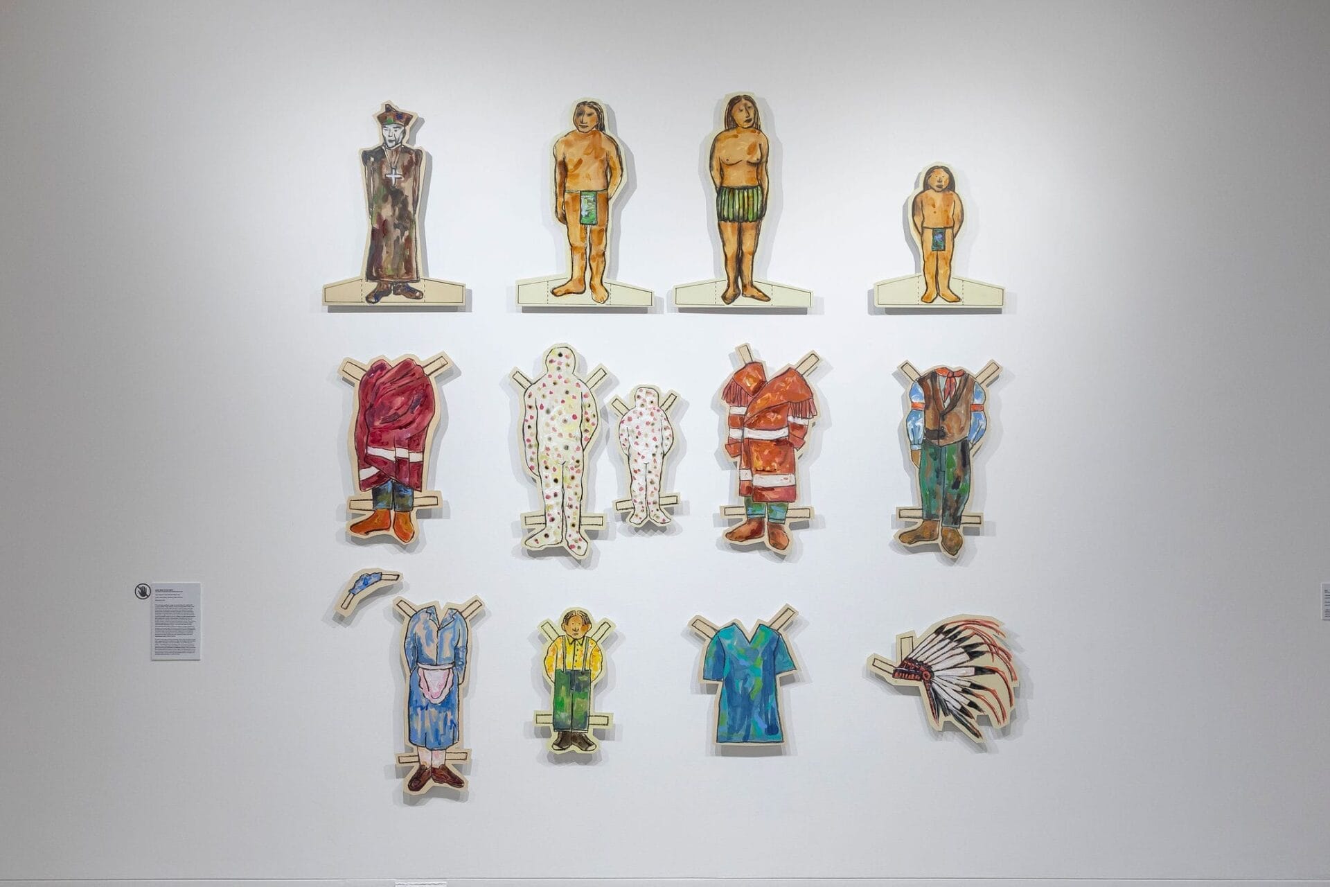 Art installation of large paper dolls crafted as Paper Cutouts with removable outfits. The top row shows four undressed figures; below are various colorful outfits and an indigenous feather headdress displayed on a white gallery wall.