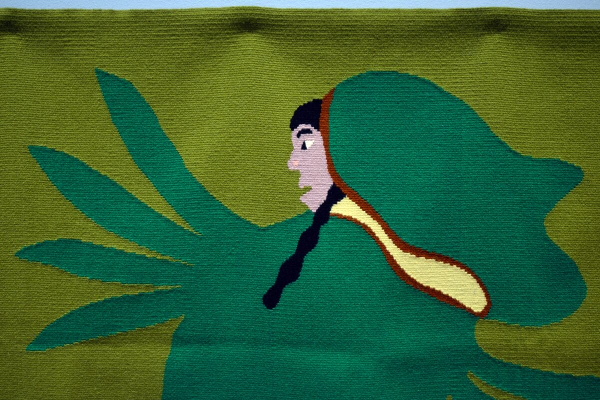 A woven textile art piece showcases visual storytelling, depicting a person in profile with long black hair and a green hooded cloak against a leafy green background, featuring stylized shapes resembling leaves or wings.