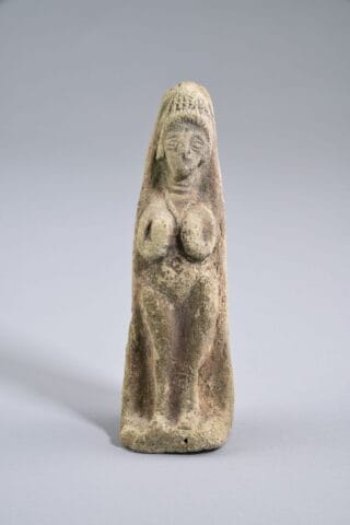An ancient stone figurine depicting a standing human figure with exaggerated features. The figure has a rounded body, prominent breasts, and a detailed headdress. The stone is weathered and has a rough texture, with a greyish-brown color.