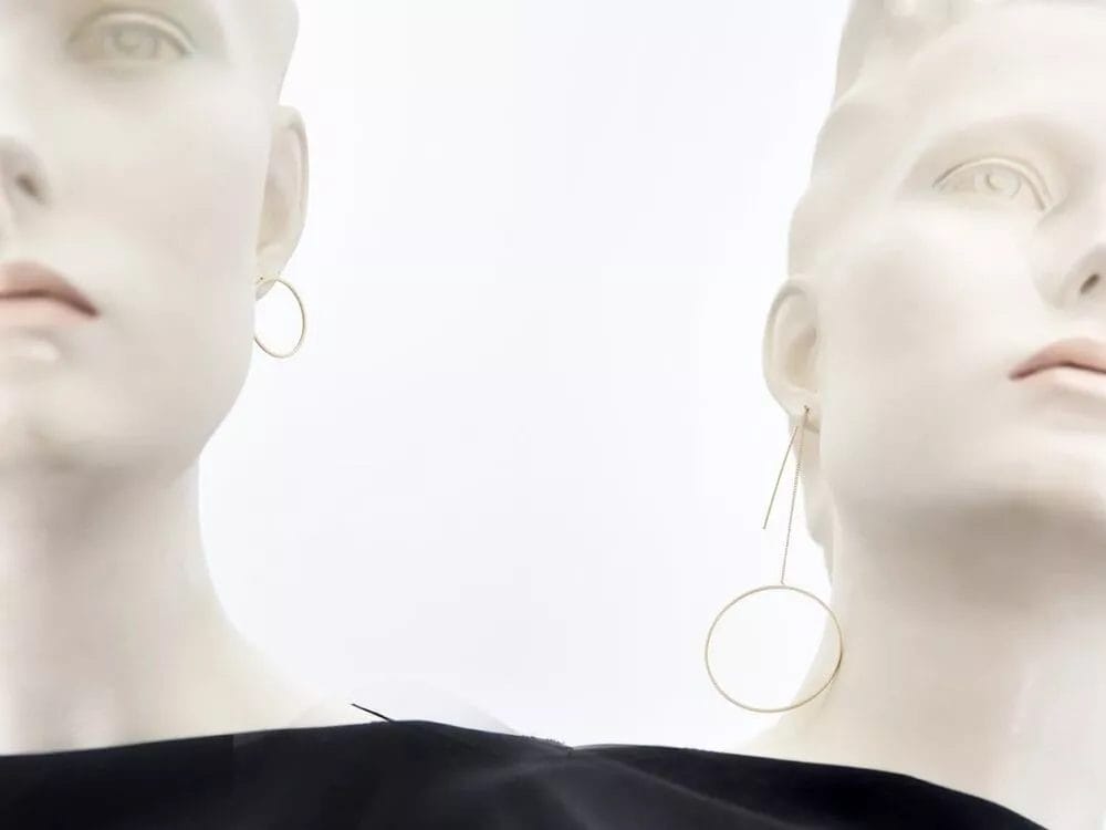 Two white mannequins with angular, spiked head designs pose in black clothing. Each wears a Pendulum Combi Earrings by Pursuits. The background is a stark white, highlighting the mannequins' minimalist, futuristic appearance and emphasizing the contrast with the earrings.
