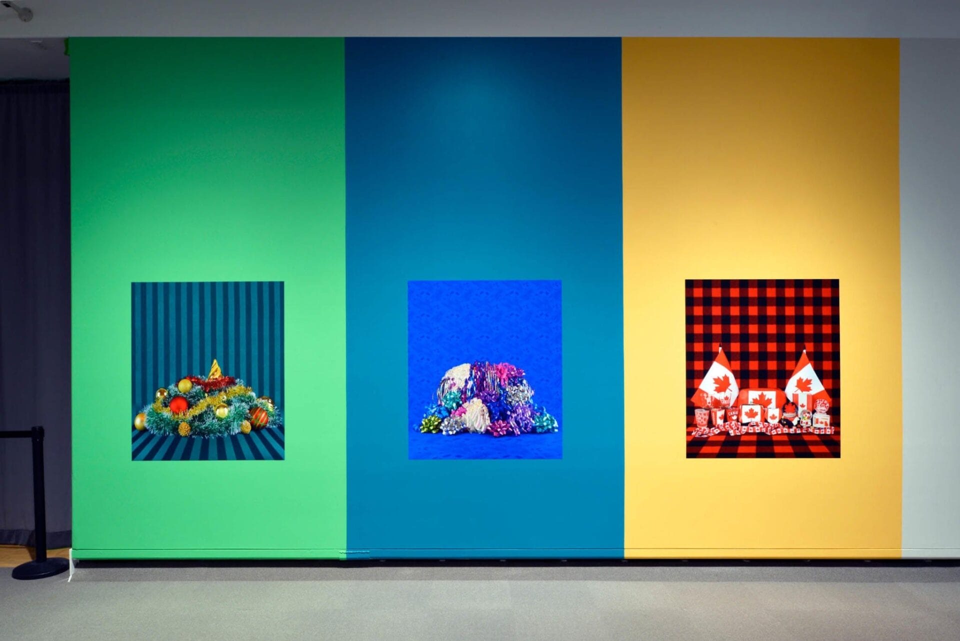 Une exposition murale colorée composée de trois panneaux. Le panneau de gauche est vert avec un fond rayé vert et bleu foncé, affichant divers fruits. Le panneau du milieu est bleu et présente des fleurs assorties sur fond bleu. Le panneau de droite est jaune avec un fond à carreaux rouge et noir, affichant deux drapeaux canadiens et une collection de petites boîtes.