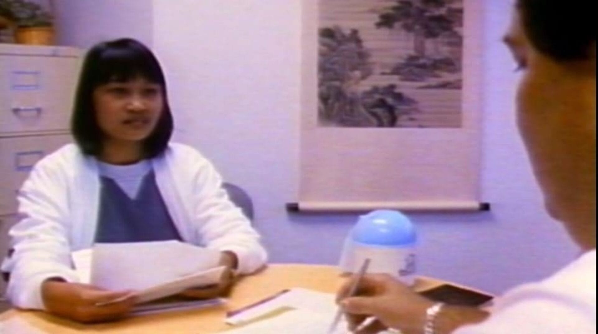 Une femme vêtue d’une blouse blanche et assise à un bureau parle à un homme, partiellement visible de dos. Elle tient des papiers et il y a un classeur et un rouleau de peinture en arrière-plan. L'homme écrit sur un cahier.