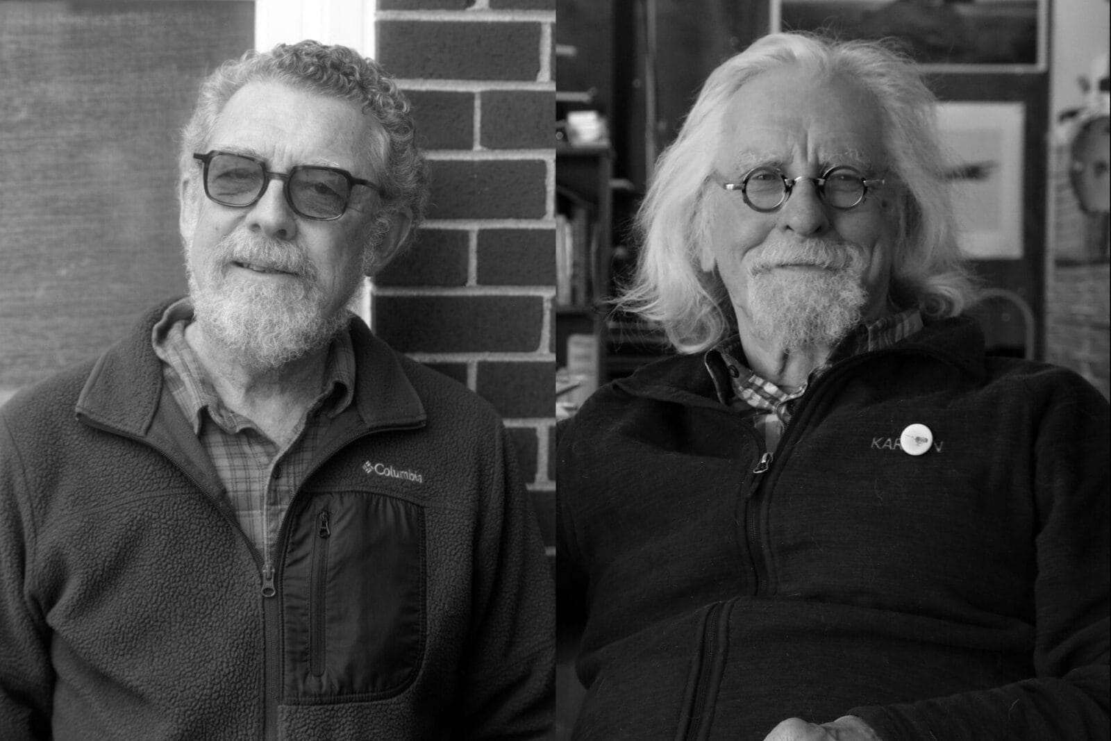 Two older men with beards and glasses sit side by side, smiling. Both wear zip-up jackets, and one has a pin on his lapel. They appear relaxed, sitting in front of a brick wall and a window. The image is in black and white.