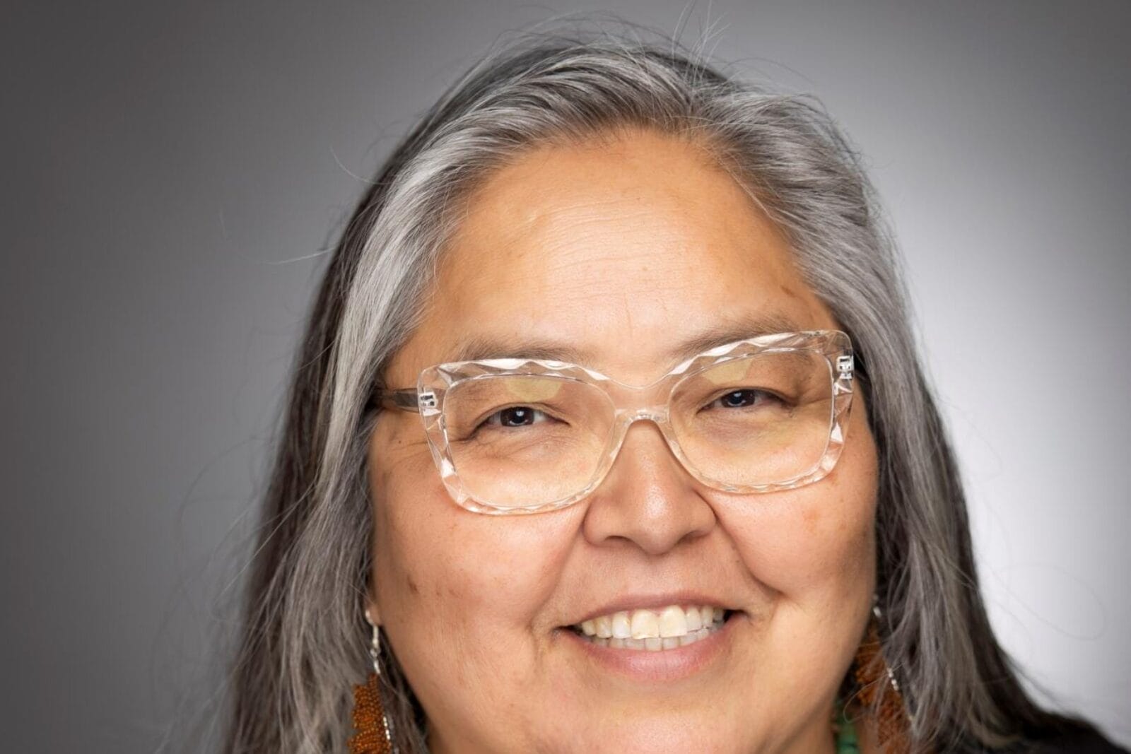 A smiling TD Curatorial Fellow with long gray hair is wearing clear glasses, a black shirt, a turquoise necklace, and beaded earrings, posing against a gray background.