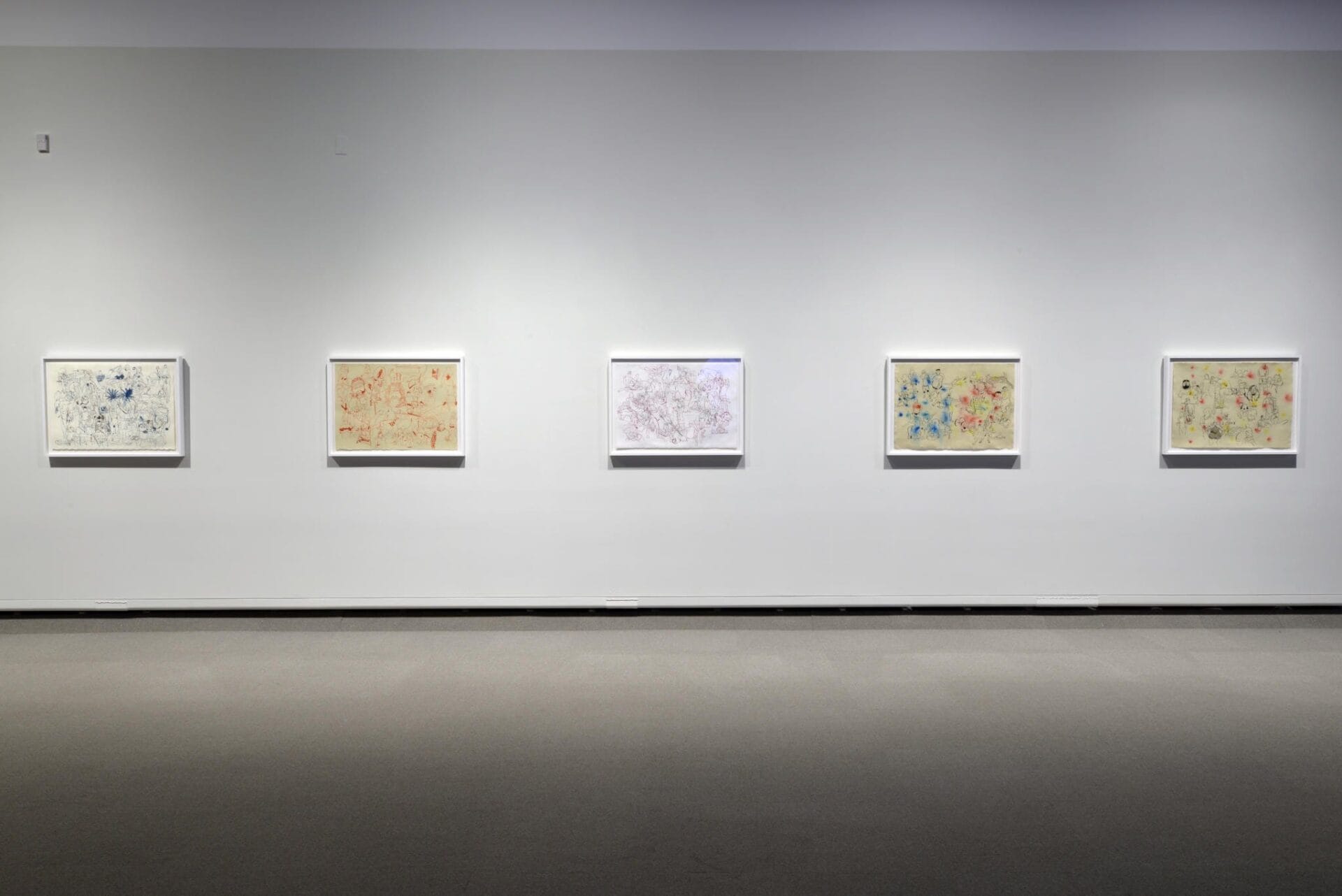 A gallery wall features five framed abstract paintings evenly spaced in a horizontal line. Each artwork showcases a colorful, intricate design with varying patterns and textures, all set against a plain, light-gray background. The floor is carpeted in gray.