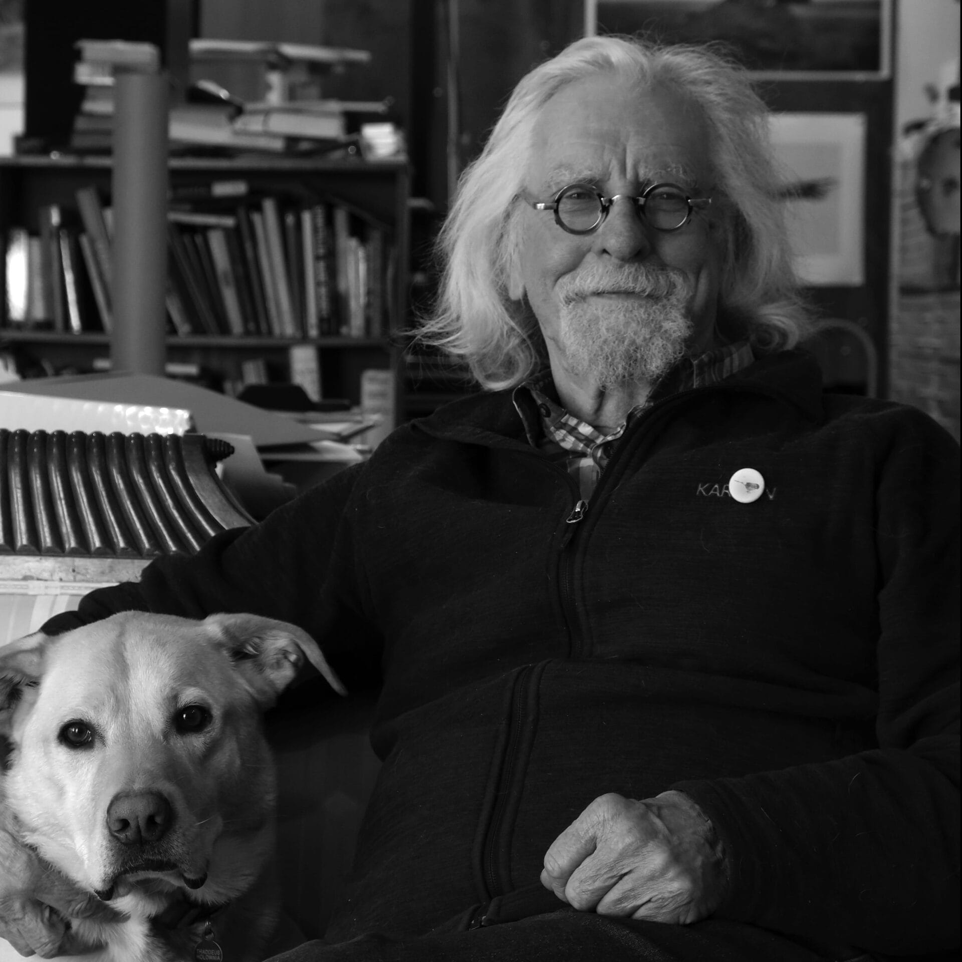 Un homme âgé aux cheveux longs et aux lunettes est assis à l'intérieur, souriant doucement, avec un chien reposant à ses côtés. Des livres et des œuvres d'art sont visibles à l'arrière-plan. L'image est en noir et blanc.