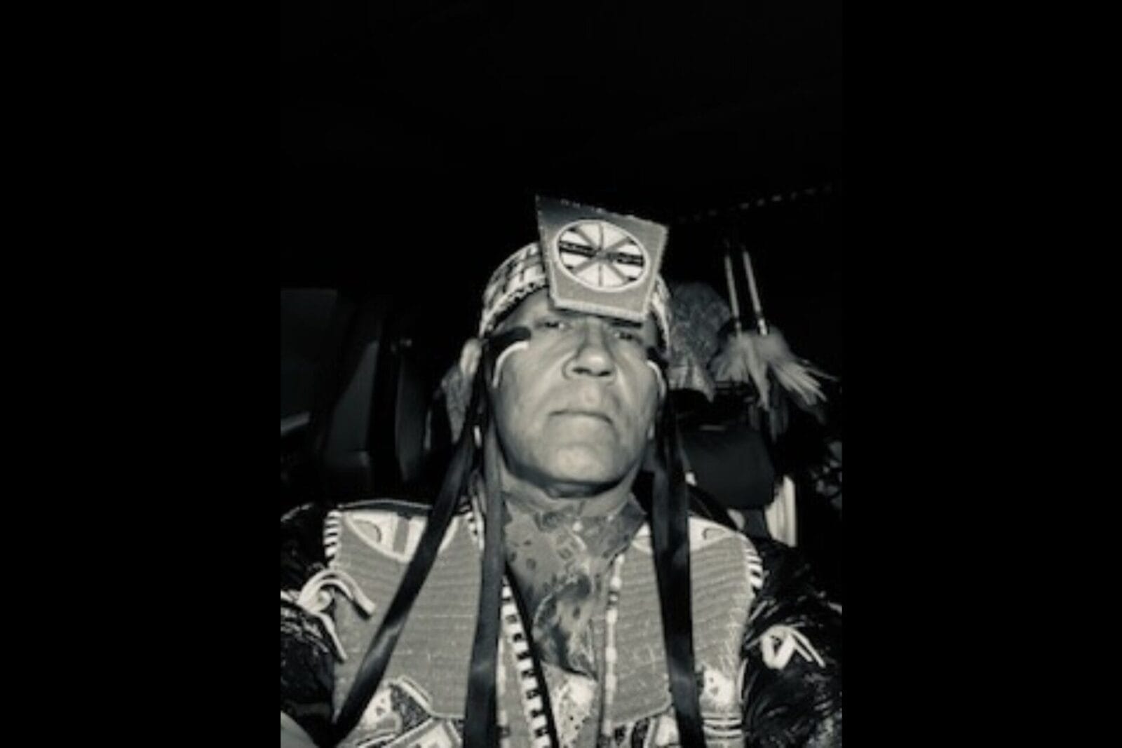 A person wearing traditional Native American regalia, including a decorated headband and outfit, looks straight at the camera in a black-and-white photo. This image is featured in Pahkwêsikan ekwa Maskihkîwâpoy Asici (Bannock and Tea) with Rod Belanger.