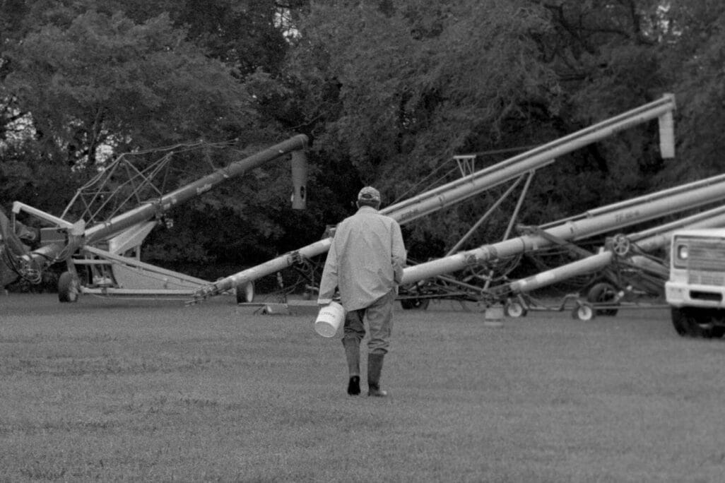 A person wearing a raincoat and boots carries a bucket while walking on grass toward large pieces of farming equipment, with trees and a truck in the background.