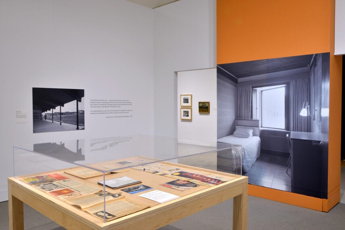Exposition muséale présentant une vitrine avec divers documents et images. L'arrière-plan montre une grande photo en noir et blanc d'un intérieur minimaliste, ainsi qu'une photo plus petite d'un bâtiment. Les murs sont principalement blancs avec une touche orange.