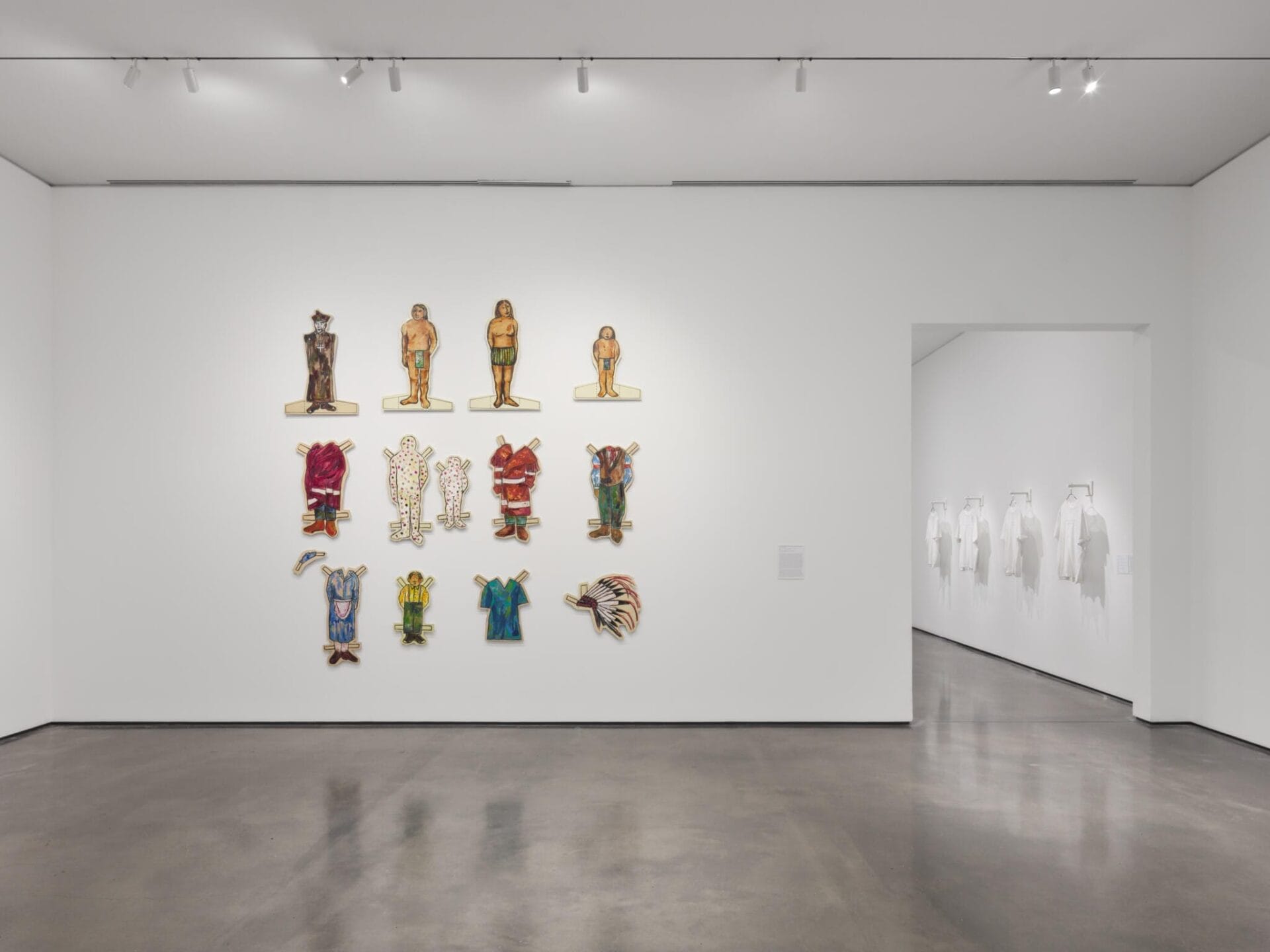 A white-walled gallery displays colorful carved figures, masks, and striking paper cutouts arranged in rows on one wall; in the next room, several white garments hang on the walls. The polished concrete floor gleams under bright lighting.