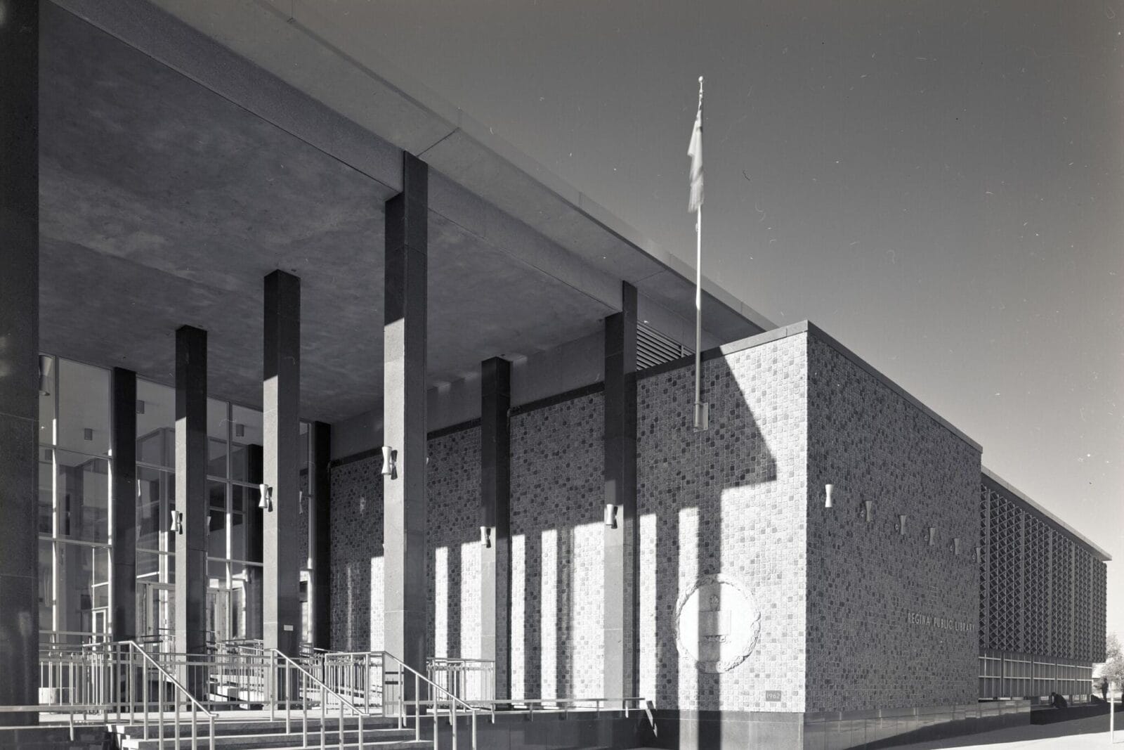 Black and white image of a modernist-style public building with large vertical pillars, a flat roof, and a brick exterior. The structure features a prominent entrance with a glass facade, multiple steps, and handrails. A flagpole is visible near the entrance, adding to the serene atmosphere reminiscent of Spring on the Prairies.